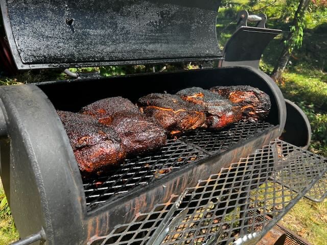 Five pork shoulders smoking on the racks of a long offset barrel smoker, with green trees behind it