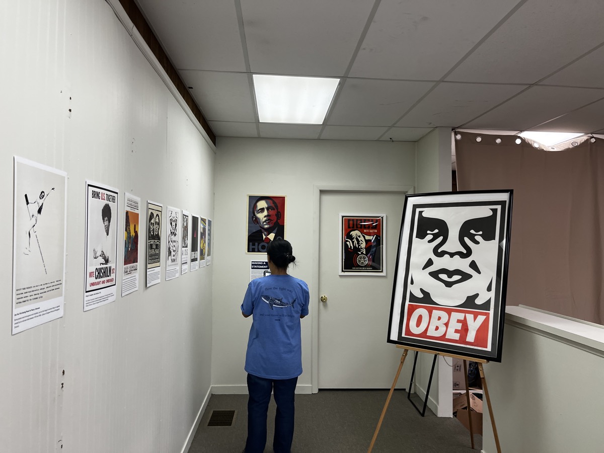 A visitor in a blue shirt looking at a row of printed political posters along the headquarters hallway, with the Obama Hope portrait and a framed poster on an easel further down