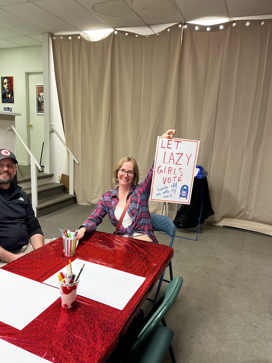 A woman at the make-your-own table smiling and holding up a hand-lettered poster reading Let Lazy Girls Vote, hands off our vote by mail, with cups of markers and blank poster board in front of her