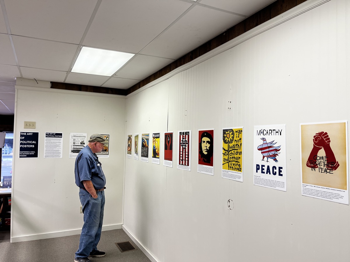 A visitor in a ball cap and blue shirt studying the exhibit wall, hung with peace and campaign posters beside the printed exhibit title cards