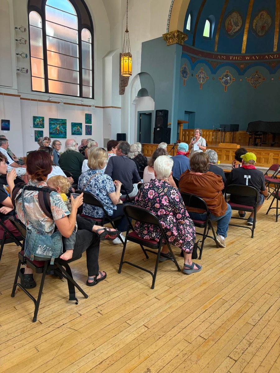 An audience seated in folding chairs in the Arts Center hall as a host introduces the performance