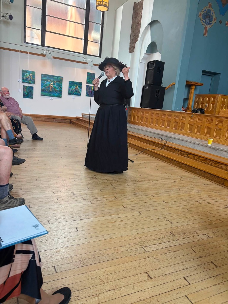 Karen Vuranch in character as Mother Jones, in a long black dress and veiled hat, speaking into a microphone in the Arts Center hall