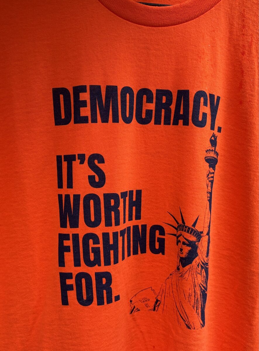 Orange T-shirt with navy print reading Democracy. It's worth fighting for, beside the Statue of Liberty